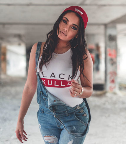 woman in white black skullz bodysuit with logo and red brand logo hat wearing overalls