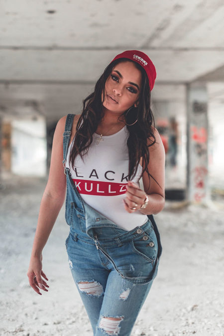 woman in white black skullz bodysuit with logo and red brand logo hat wearing overalls